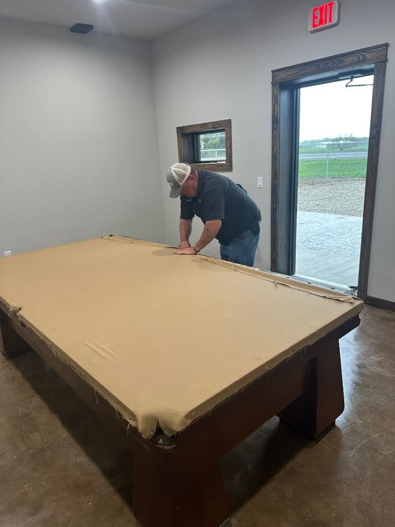 Man installing tan pool table felt in a room with a concrete floor, doorway, and an exit sign.