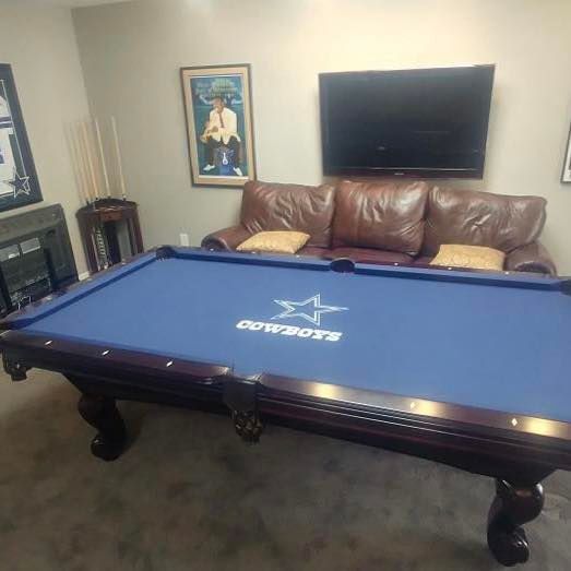 Blue pool table with Dallas Cowboys logo, brown leather couch, TV, fireplace, and sports memorabilia.