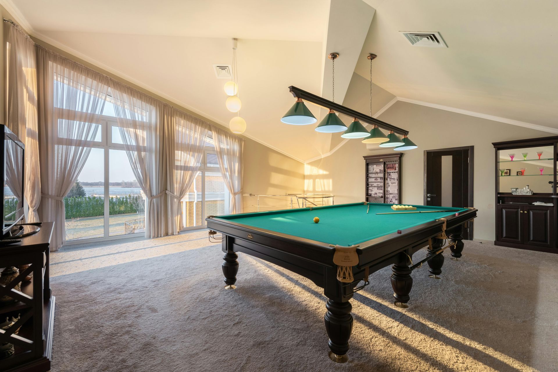 A pool table in a well-lit room with large windows, hanging lights, and a cabinet.