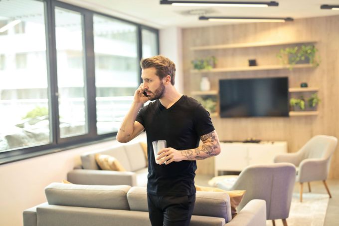 Man standing in a bright living room, talking on a phone and holding a cup.