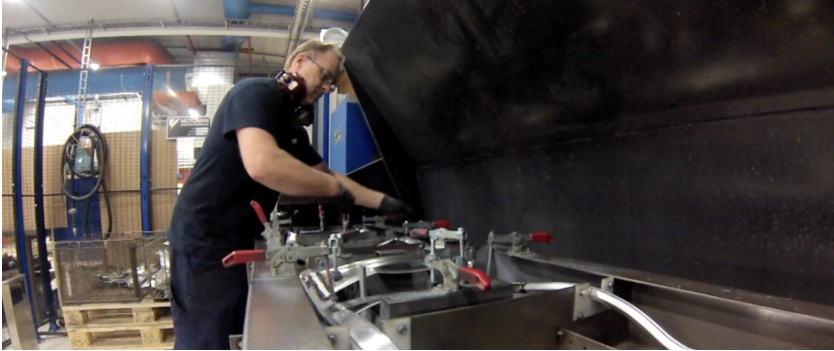 En person i et verksted i svart uniform arbeider med metallmaskiner.