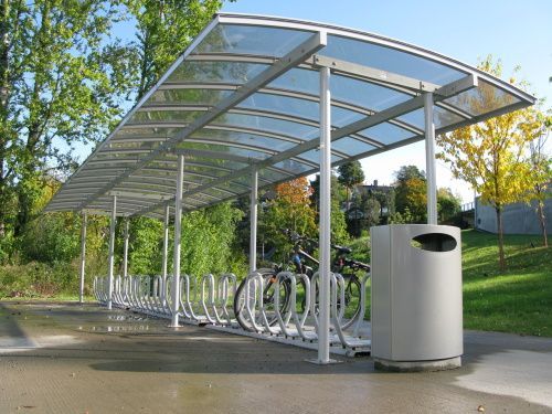 Sykkelskur med flere sykkelstativer og en søppelbøtte. Utendørs, solskinnsdag.