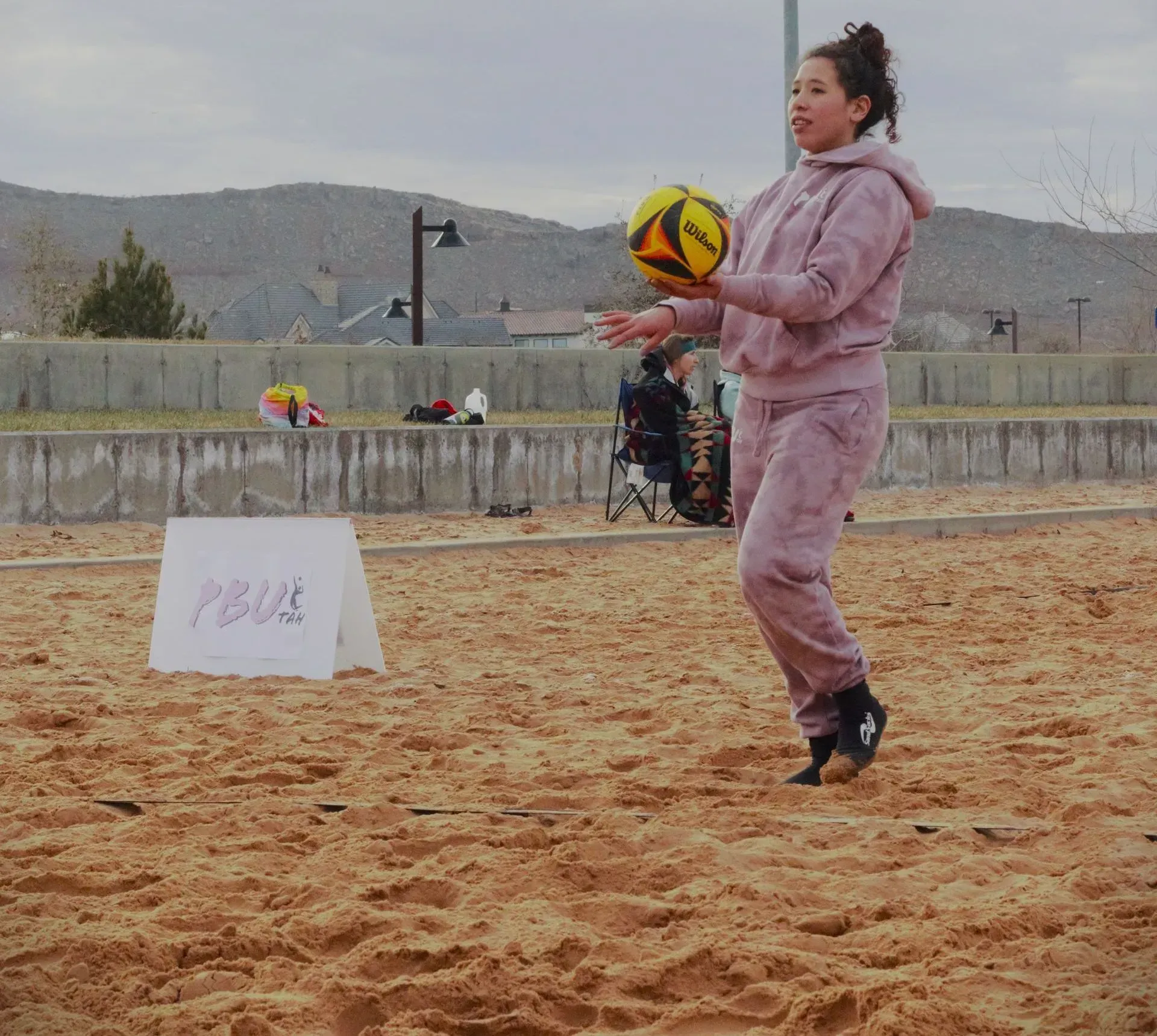 Woman playing with a volleyball on sand, wearing a pink sweatsuit. 