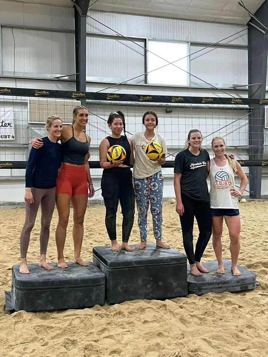Six women pose on podiums holding volleyballs on a sand court. Six women pose on podiums holding volleyballs on a sand court.