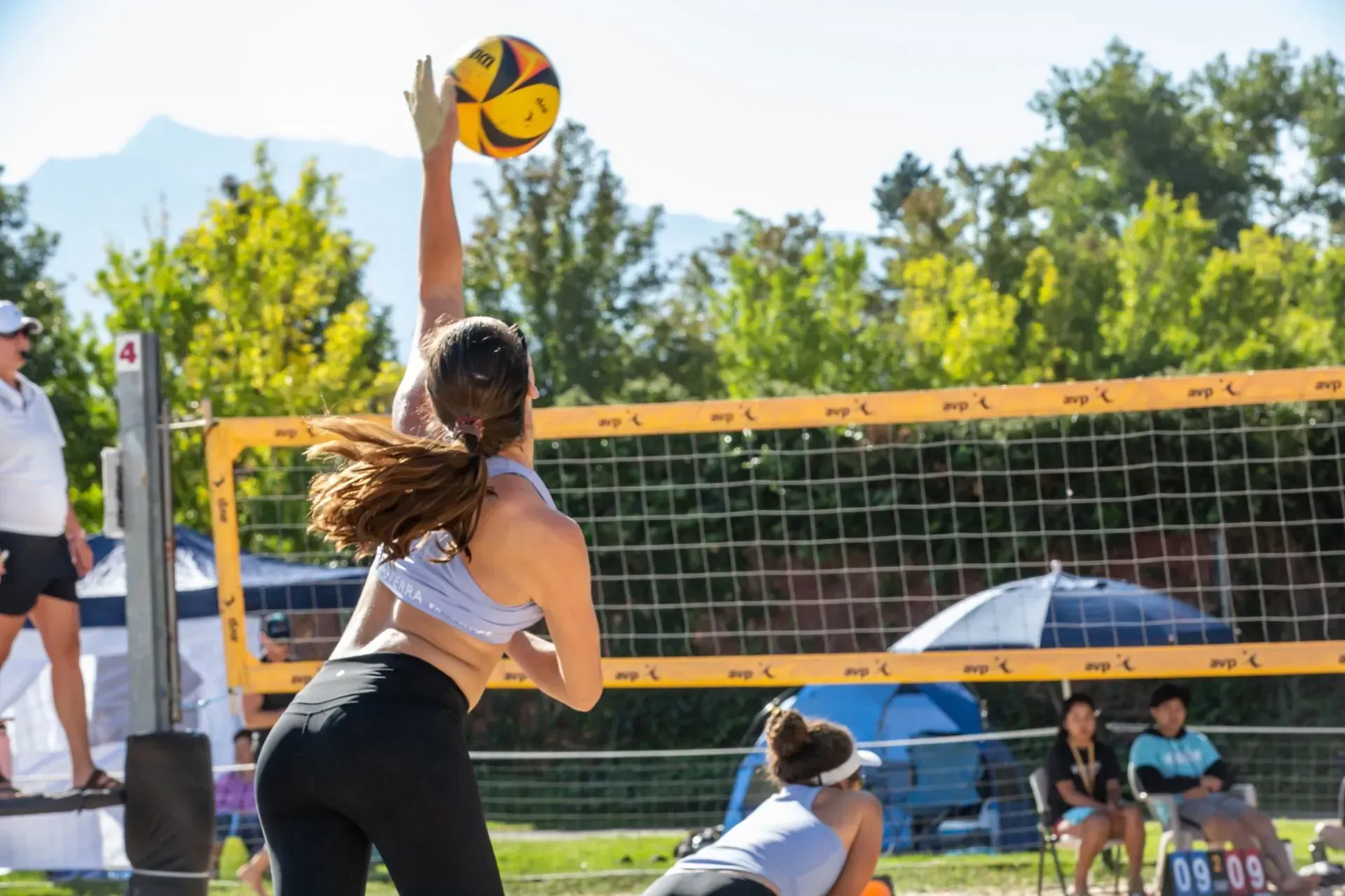 Woman in sports bra spiking volleyball on beach, net and crowd visible.
