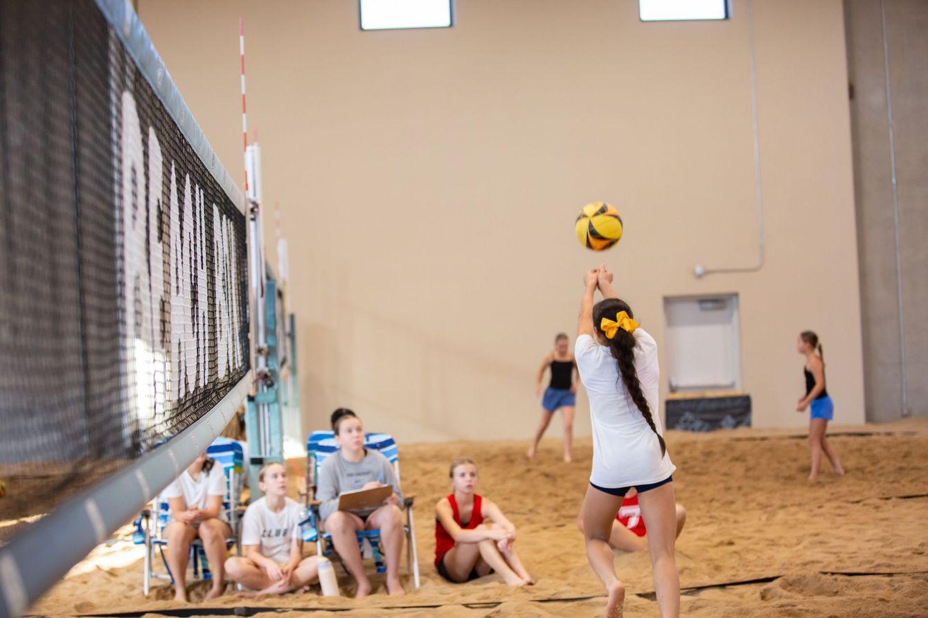 Beach volleyball game: players, net, sandy court, mountains in background, sunny day.