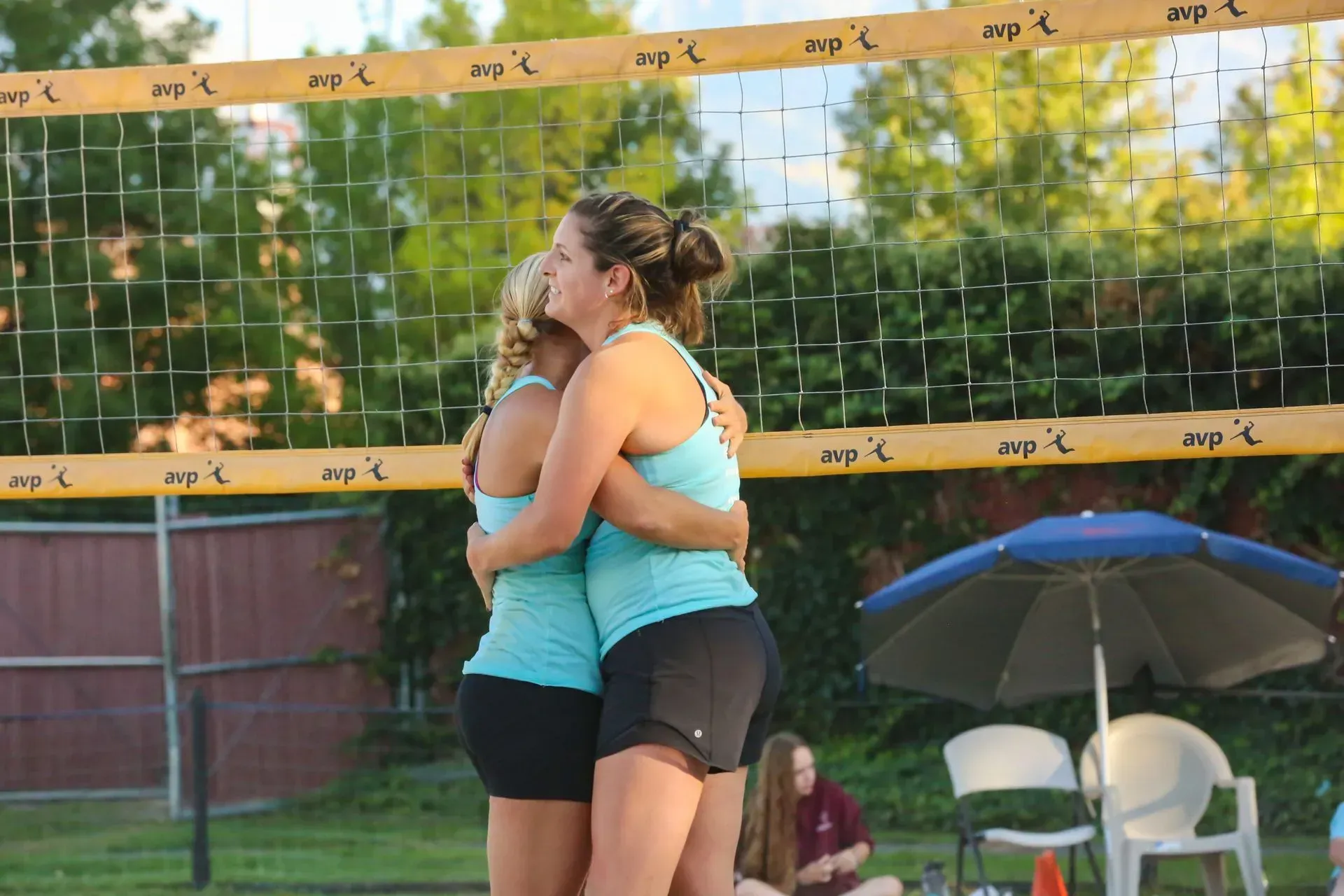 Two volleyball players in teal tank tops and shorts embrace at the net.