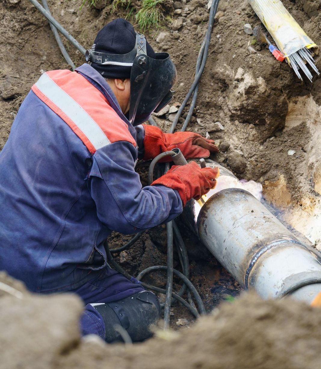 A worker in a blue uniform and welding mask welds a metal pipe in a trench; red gloves and sparks are visible.
