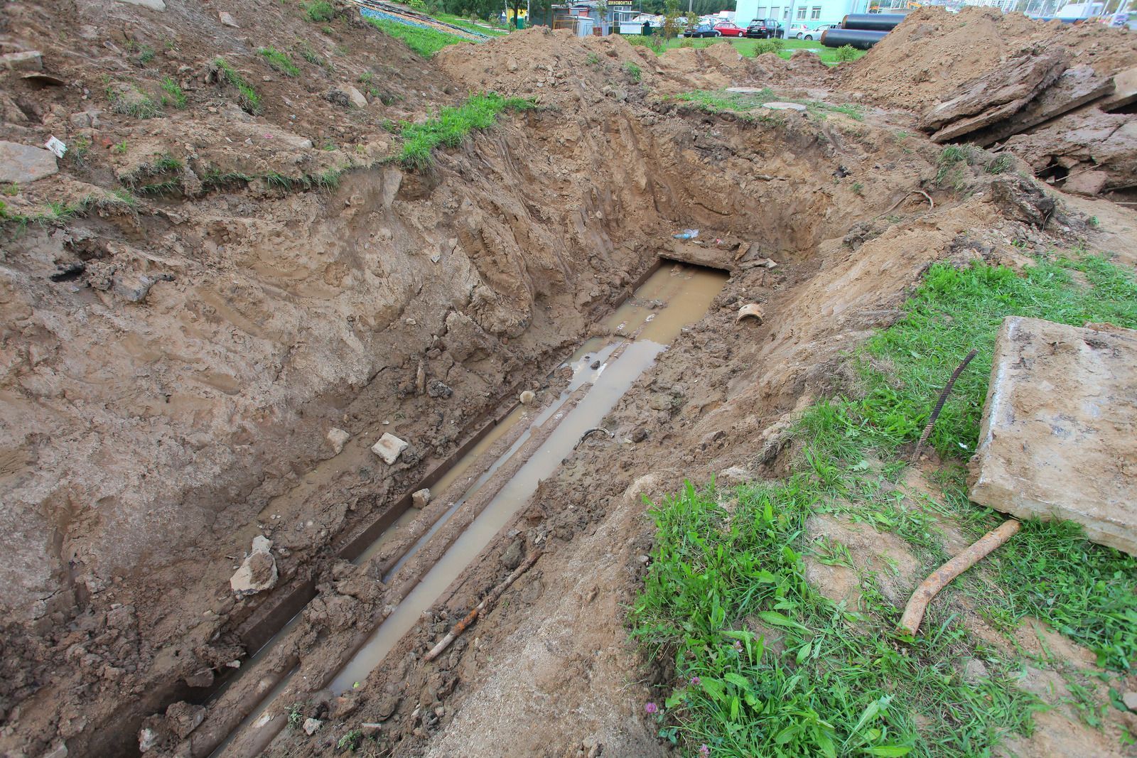 A long, open trench filled with muddy water, revealing buried pipes in a construction site.