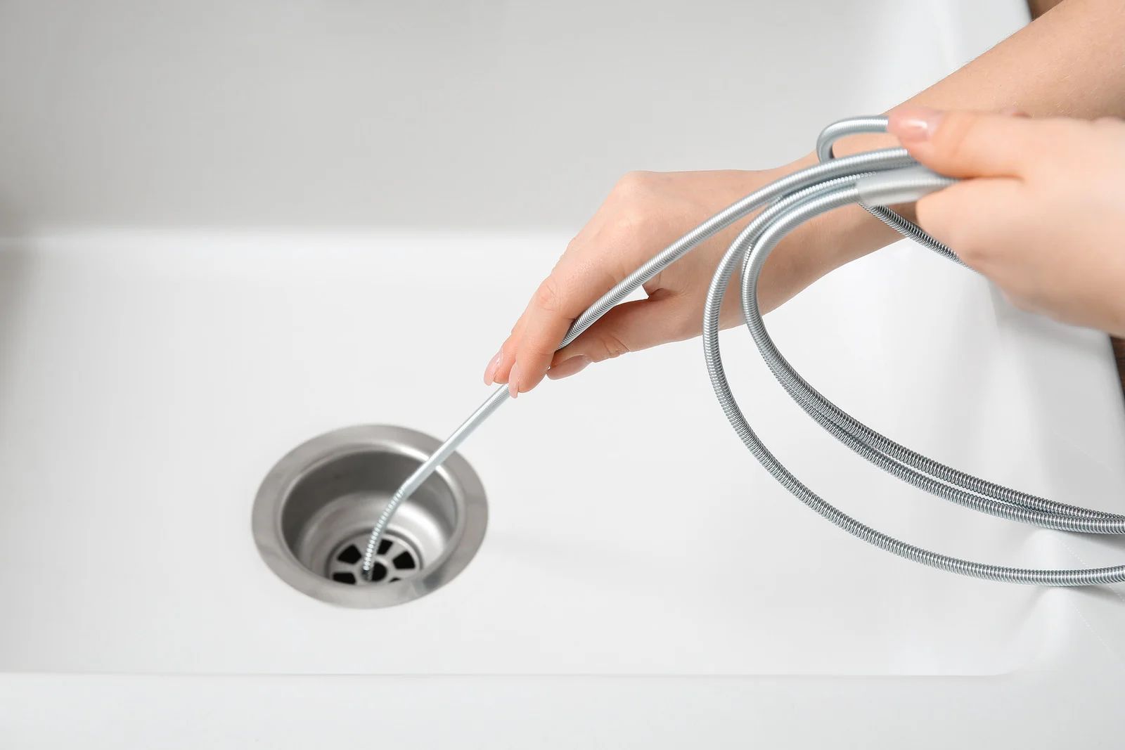 A person holds a silver drain cleaning tool and inserts it into a clean white kitchen sink drain.
