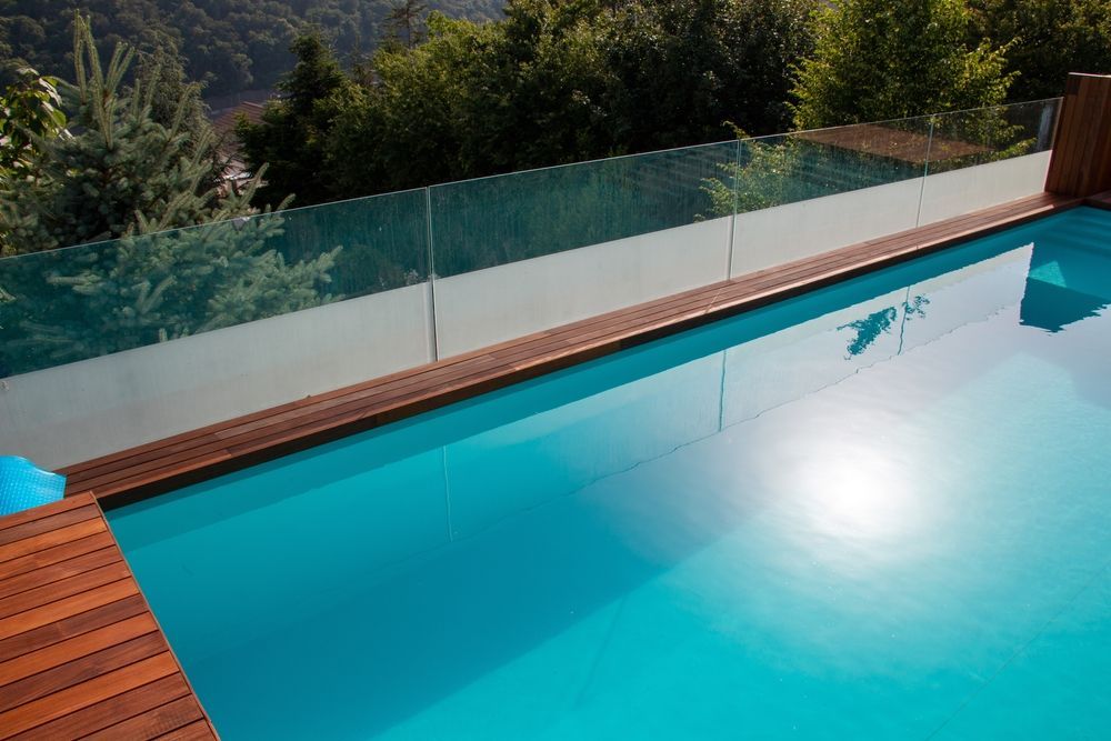 A Turquoise Swimming Pool With Wooden Decking and Glass Railing — Gympie Fence Inspections in Glenwood, QLD