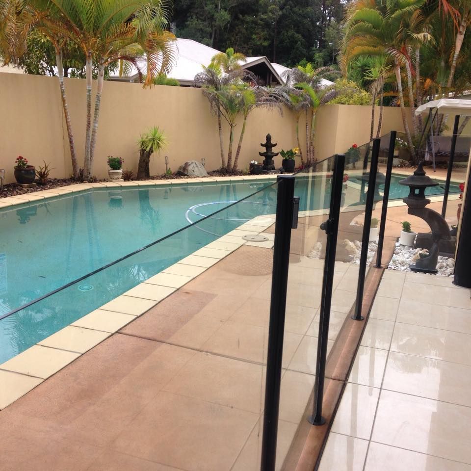 Swimming Pool With Glass Fence, Surrounded by Tan Concrete and Lush Greenery — Gympie Fence Inspections in Gympie, QLD