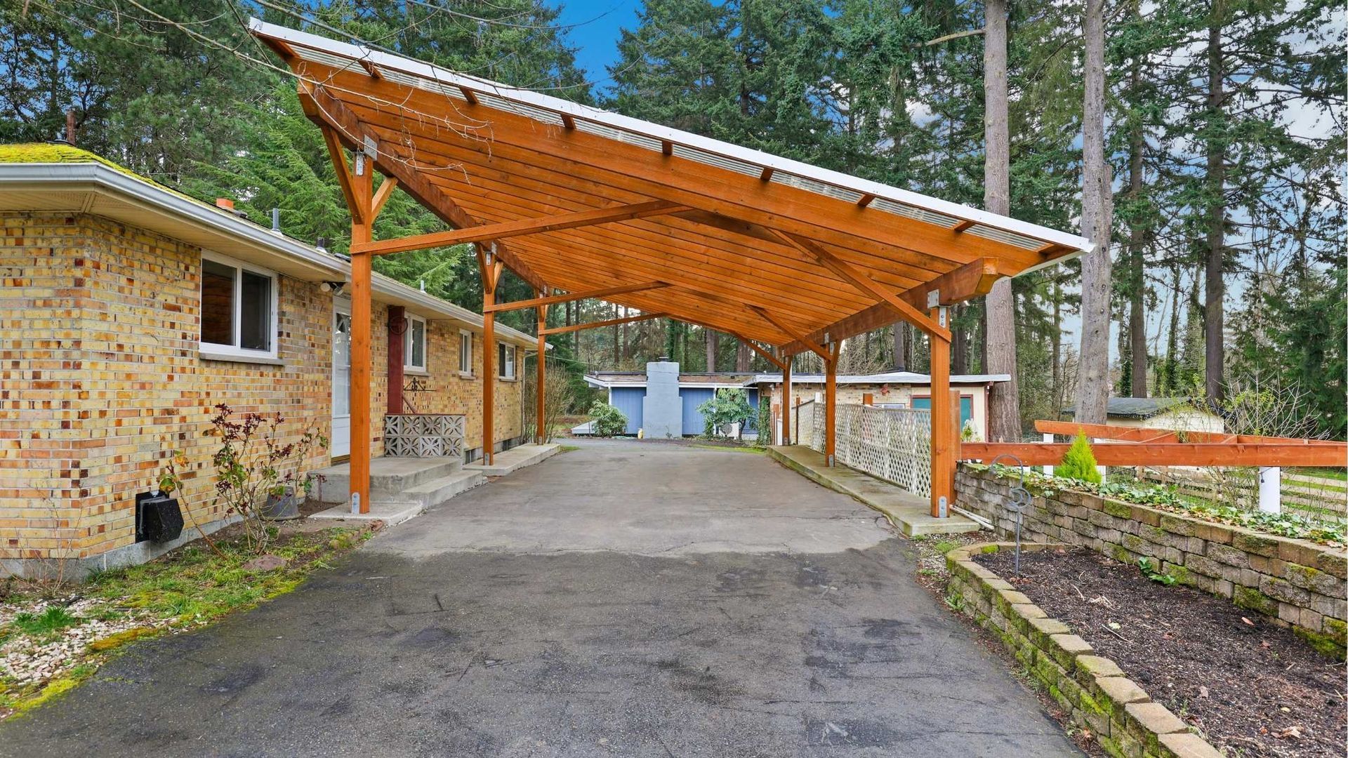 Covered driveway beside brick house and forest, with wooden carport and paved entrance