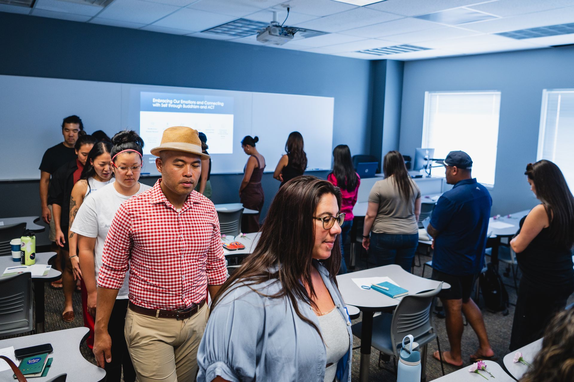 People walking in a classroom, some looking at a projector screen. Neutral expressions, bright room.