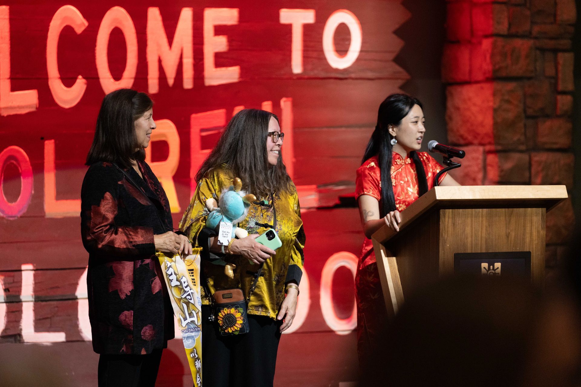 Three women on stage at an event; one speaking at a podium, two beside her holding a banner and stuffed animal.
