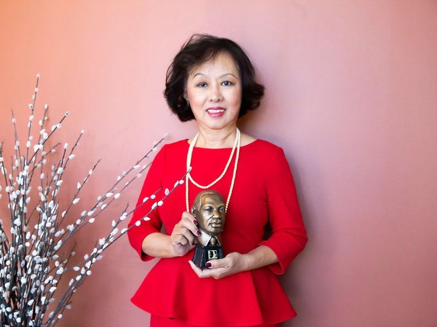 Woman in red dress holding a bust, standing in front of a peach wall, near a branch with white flowers.