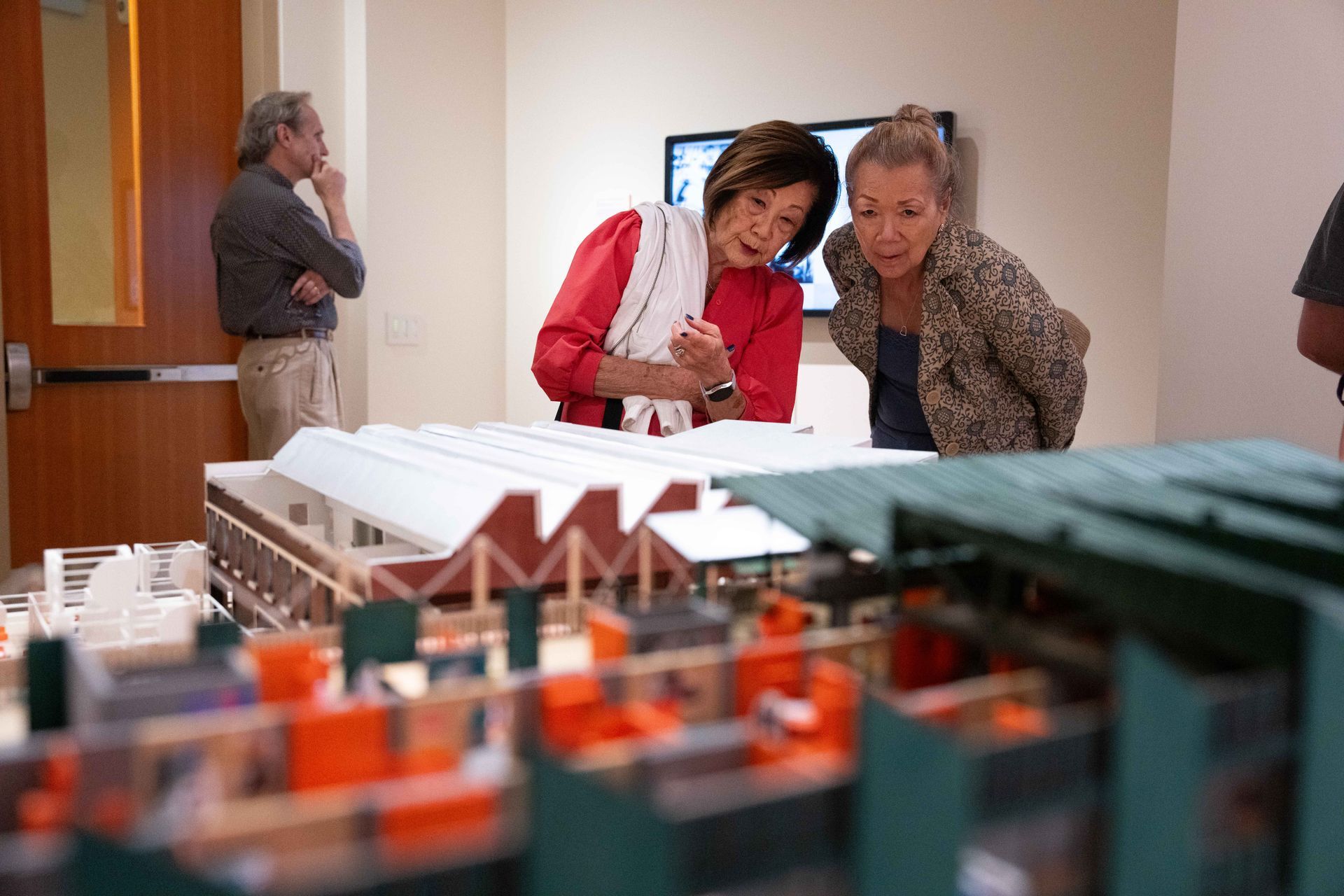 Two women examine an architectural model. A man stands nearby. A screen is in the background.