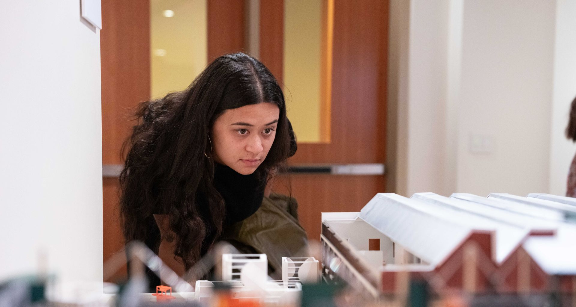 Woman with long dark hair intently examines a model building in a brightly lit room.