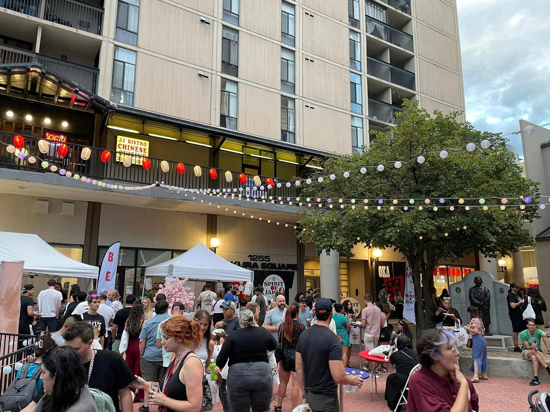 Crowd at an outdoor festival with food stalls, string lights, and a high-rise building.