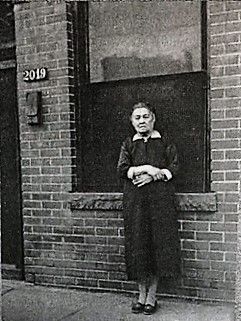 Woman standing in front of a brick building with the number 2019. She wears a long coat and looks at the camera.