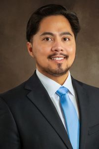 Professional headshot of a person in a dark suit and blue tie against a brown studio backdrop
