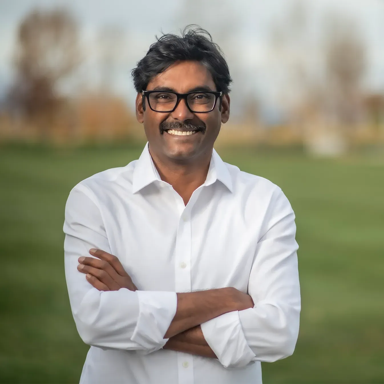 Smiling man in a white shirt with arms crossed, standing outdoors with a blurred park background