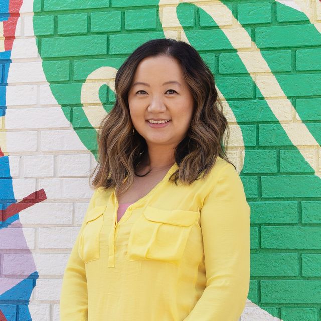 Woman with brown hair, wearing a yellow shirt, smiling in front of a colorful brick mural.