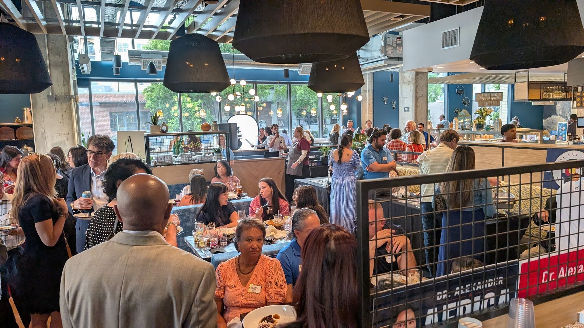 Busy café with many people seated indoors near large windows and hanging pendant lights