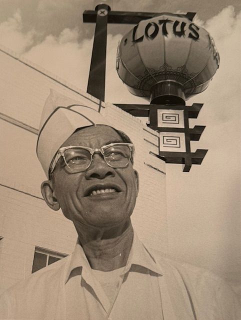 Smiling Asian man wearing glasses and a uniform stands under a Lotus restaurant sign.