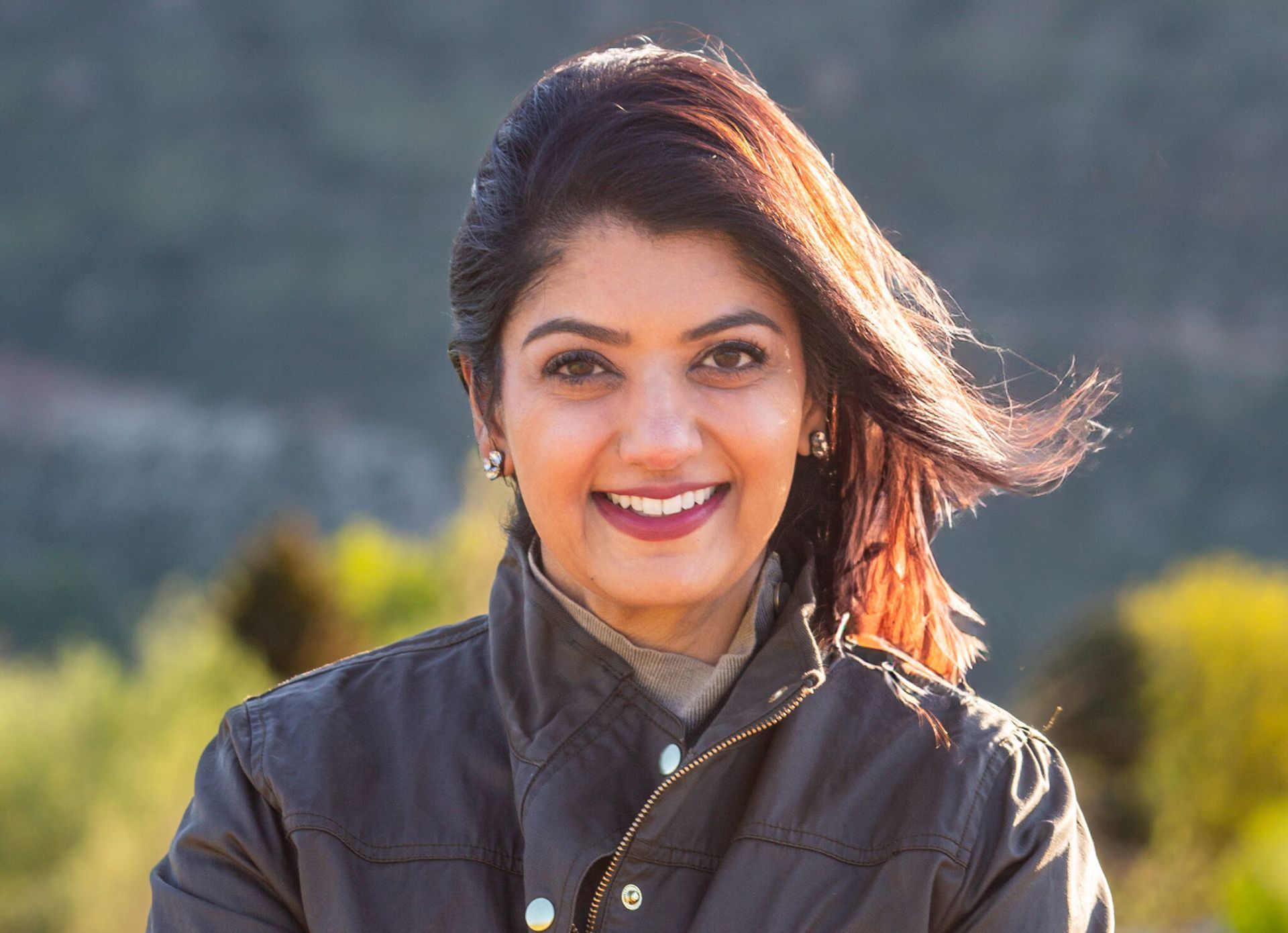 Portrait of a smiling person outdoors, wearing a dark jacket with windblown hair