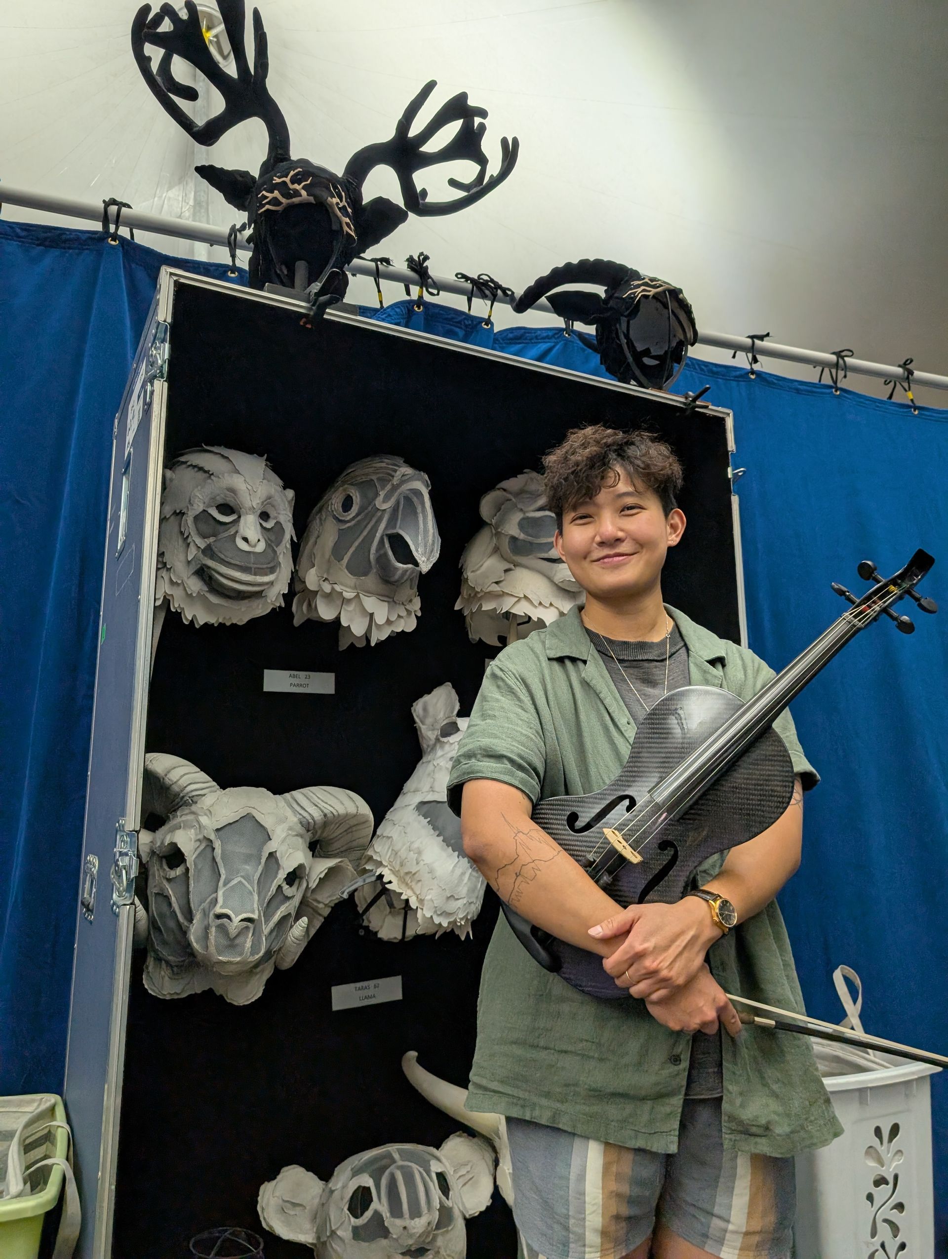 Person holding violin, smiling, posing in front of display with animal masks and antlers.