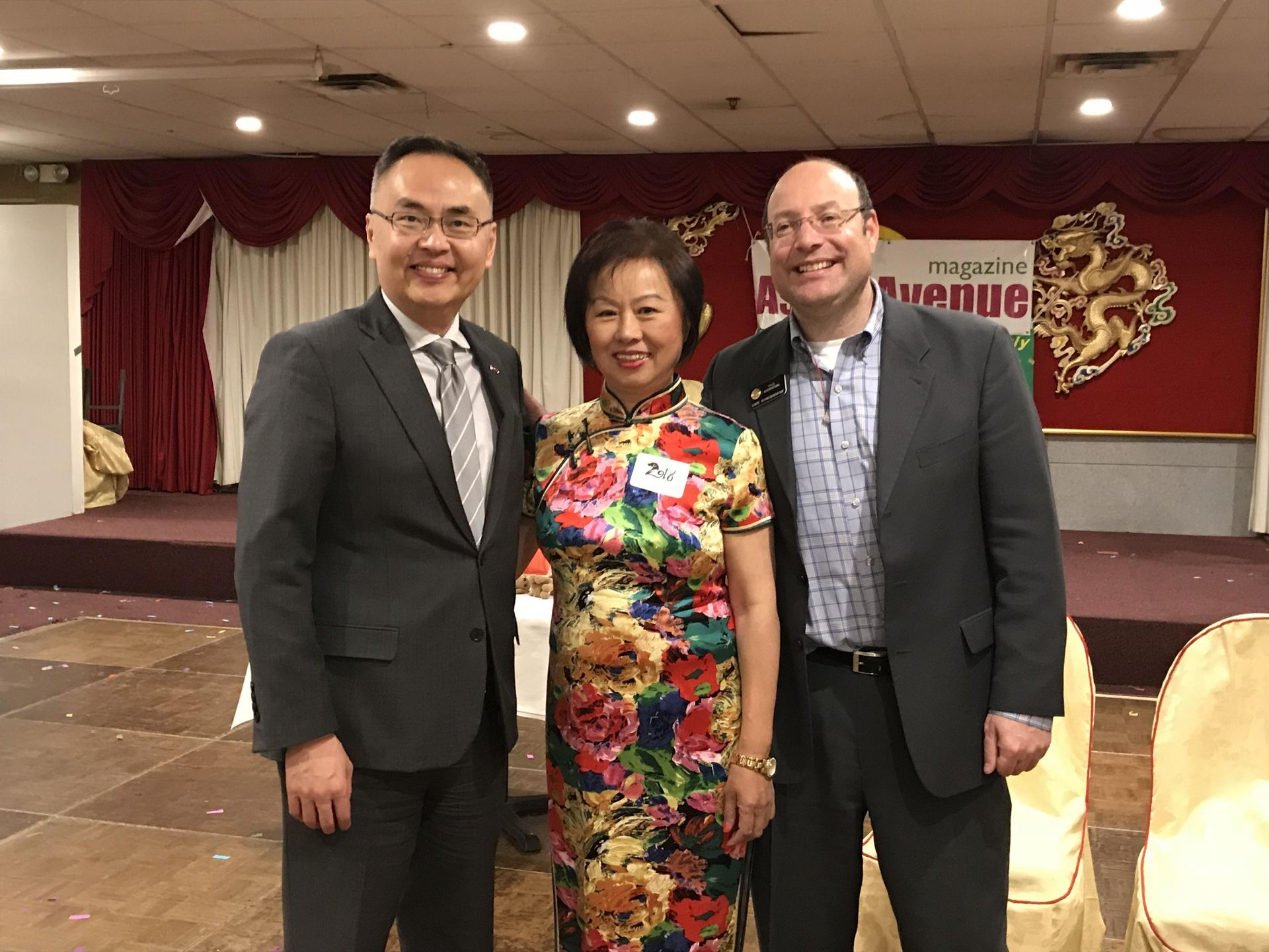 Three people posing for a photo indoors: a woman in a floral dress smiles between two men in suits.