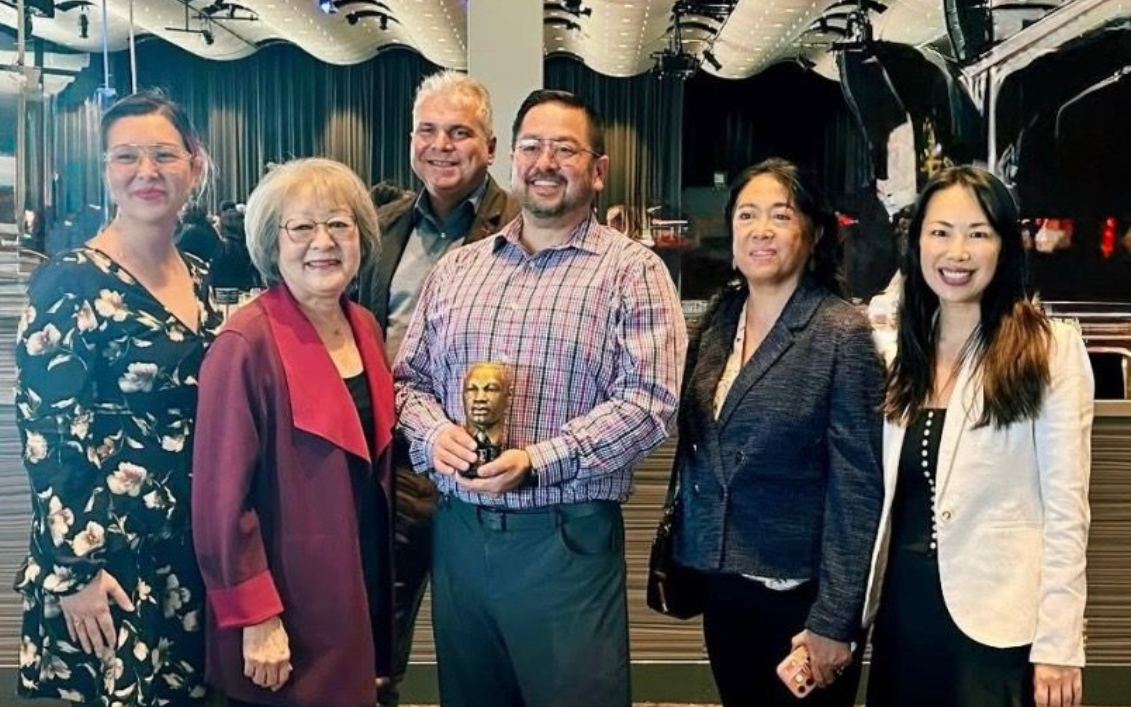 Group of people holding a trophy, indoors. Smiles, formal attire. Dark background with reflective surfaces.