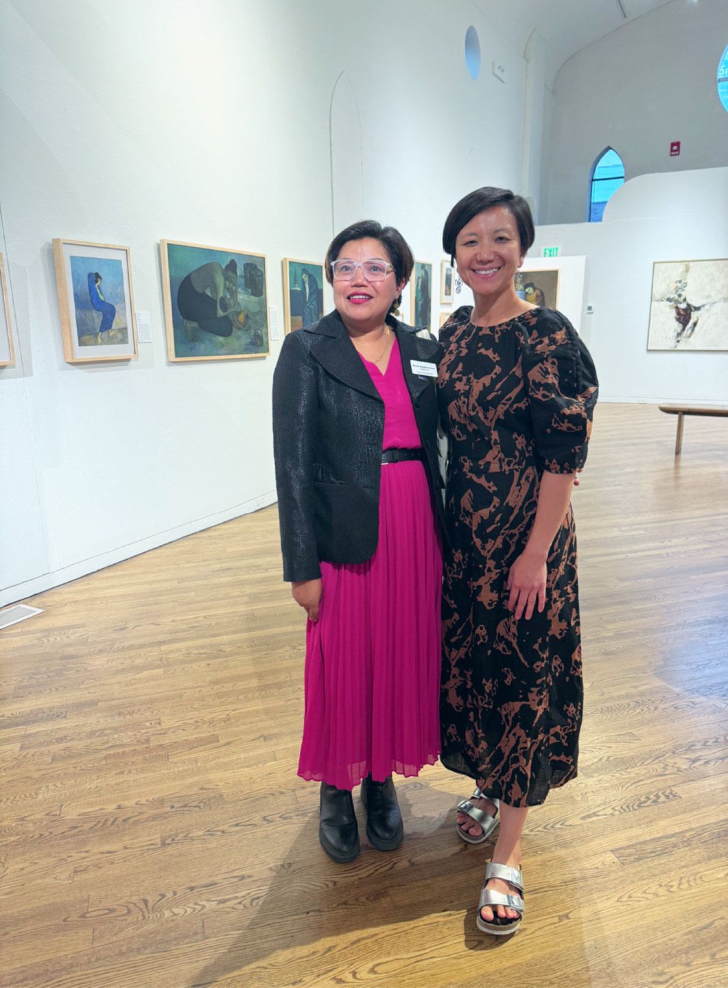 Two women pose in an art gallery. One in a pink dress, black jacket; the other in a patterned dress, arms around each other. 