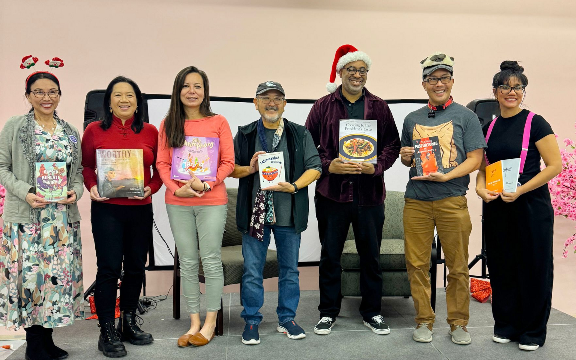 Group of people holding books at a reading event. Stage with a white backdrop, pink decor.