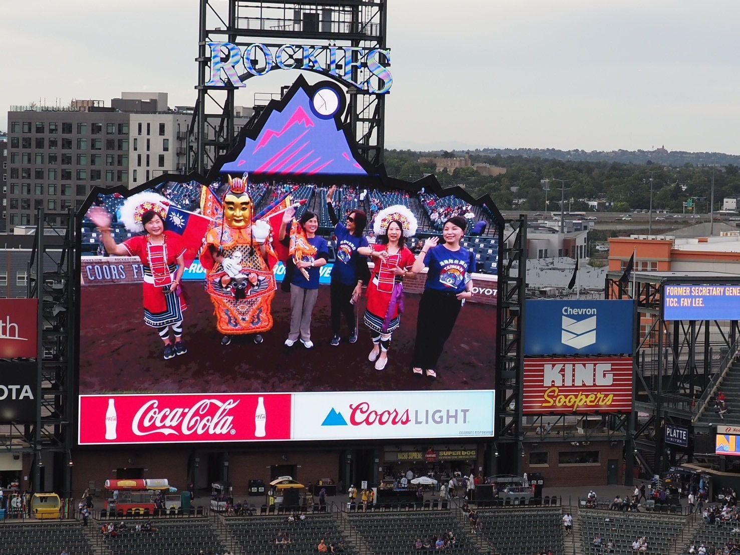 Large scoreboard at a baseball stadium shows dancers in colorful costumes and a Coca-Cola and Coors Light ad.
