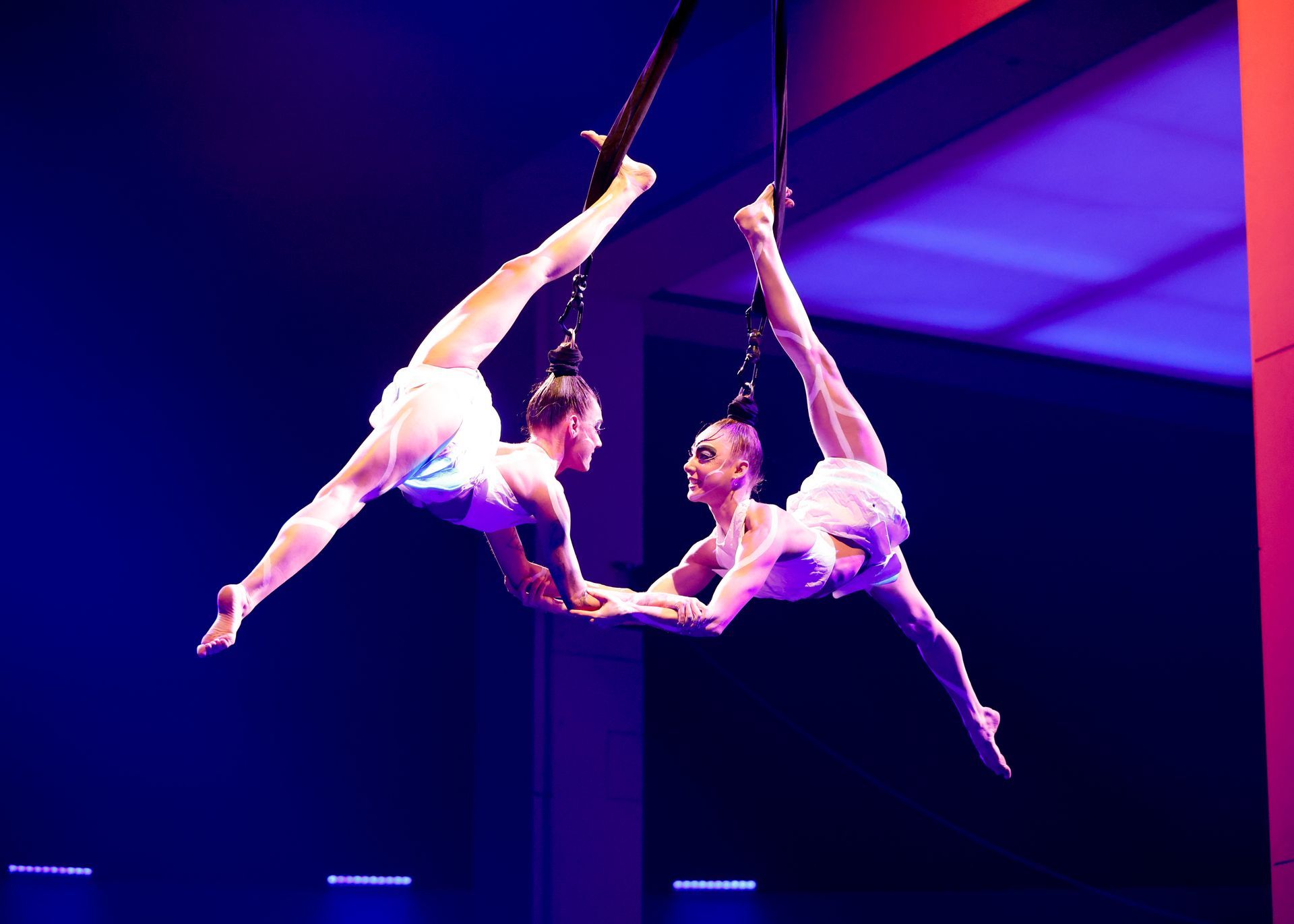 Two aerialists in white costumes perform a split in the air, facing each other.