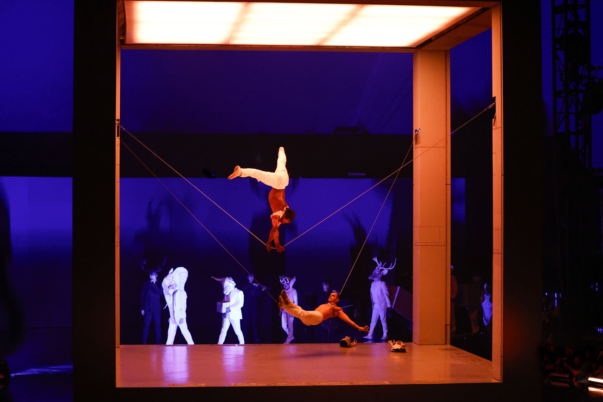 Acrobats perform inside a square-framed stage with other figures below. One acrobat is suspended upside down.