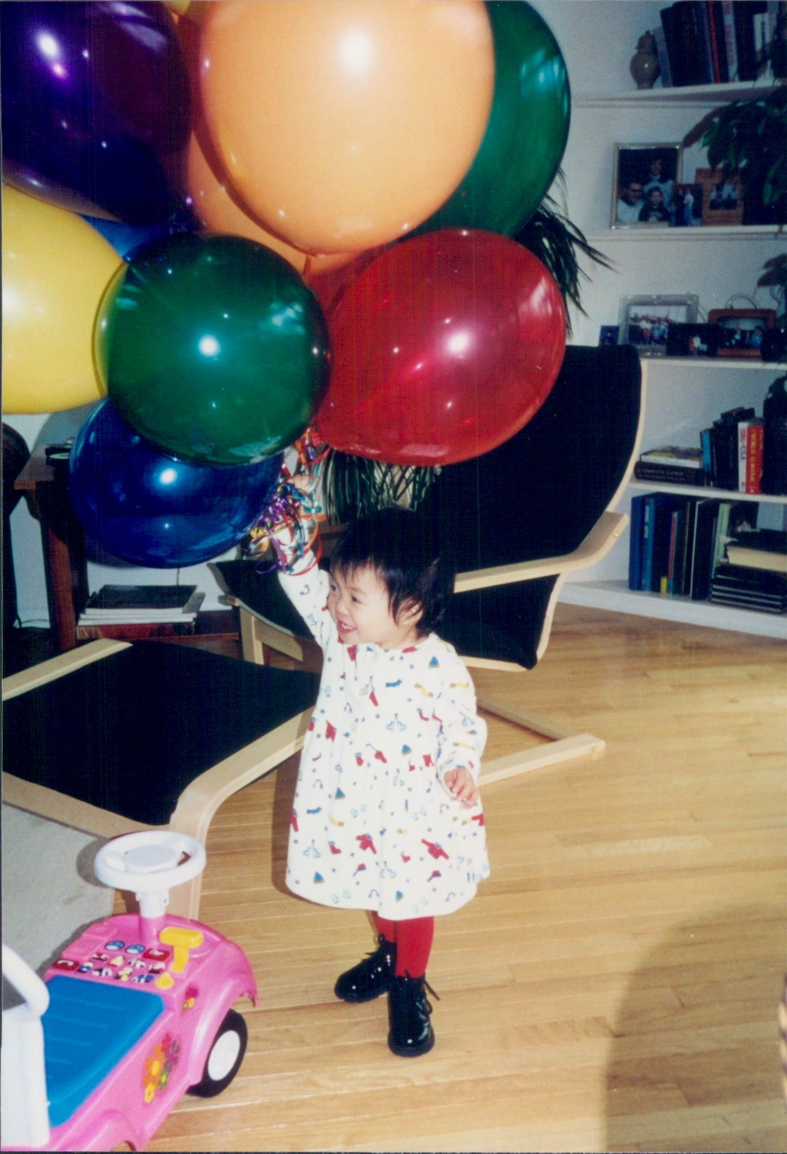 Young child in a dress holds a bunch of colorful balloons, standing by a pink toy car.