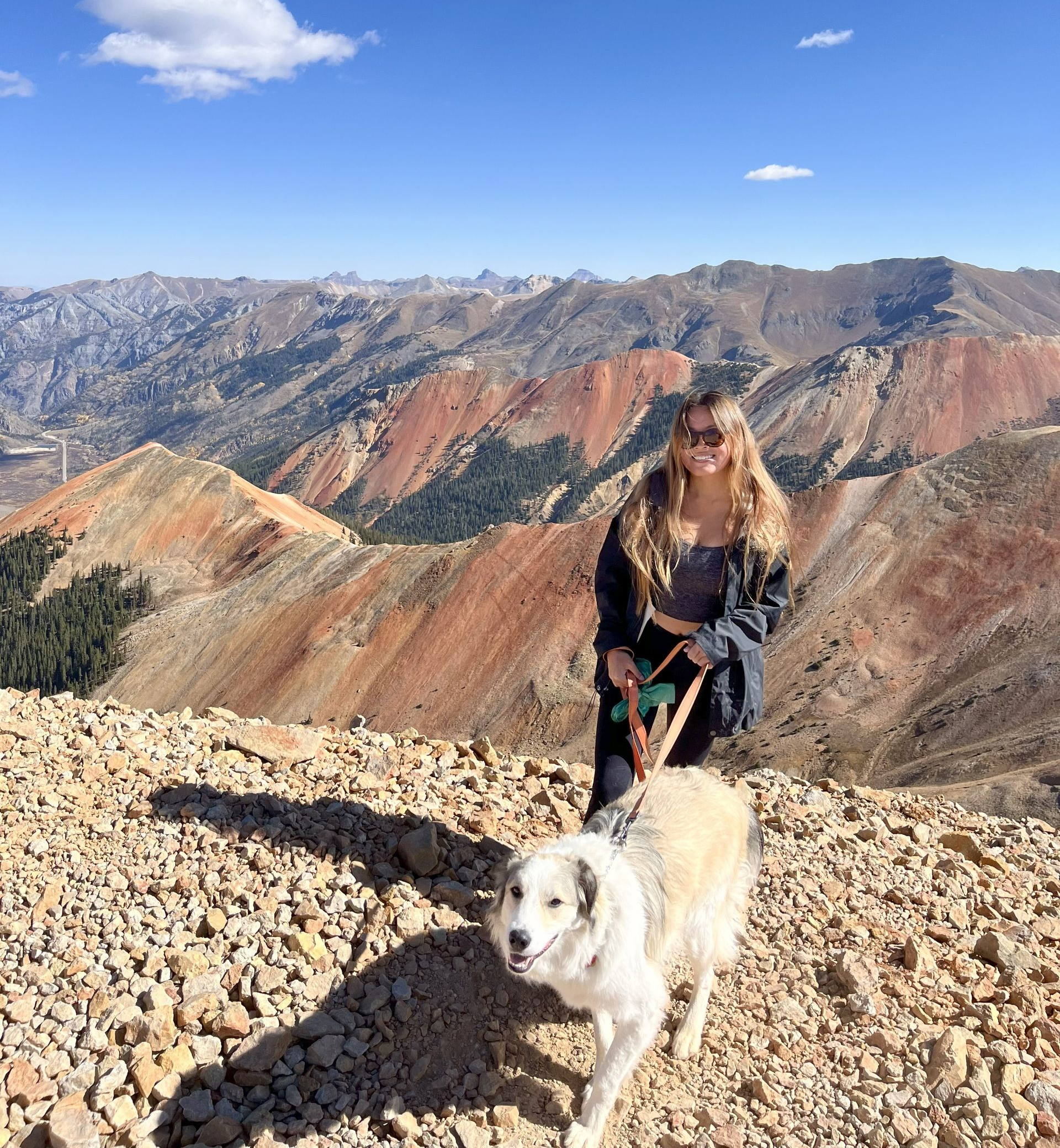 Woman and dog hiking on a rocky mountain, with a vast landscape of red and green mountains in the background.