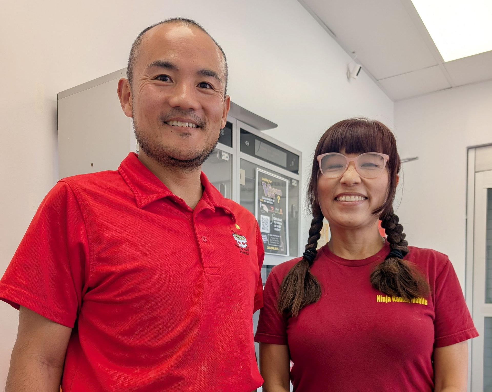 Two people smiling; man in red shirt, woman in red shirt with braids, indoors.