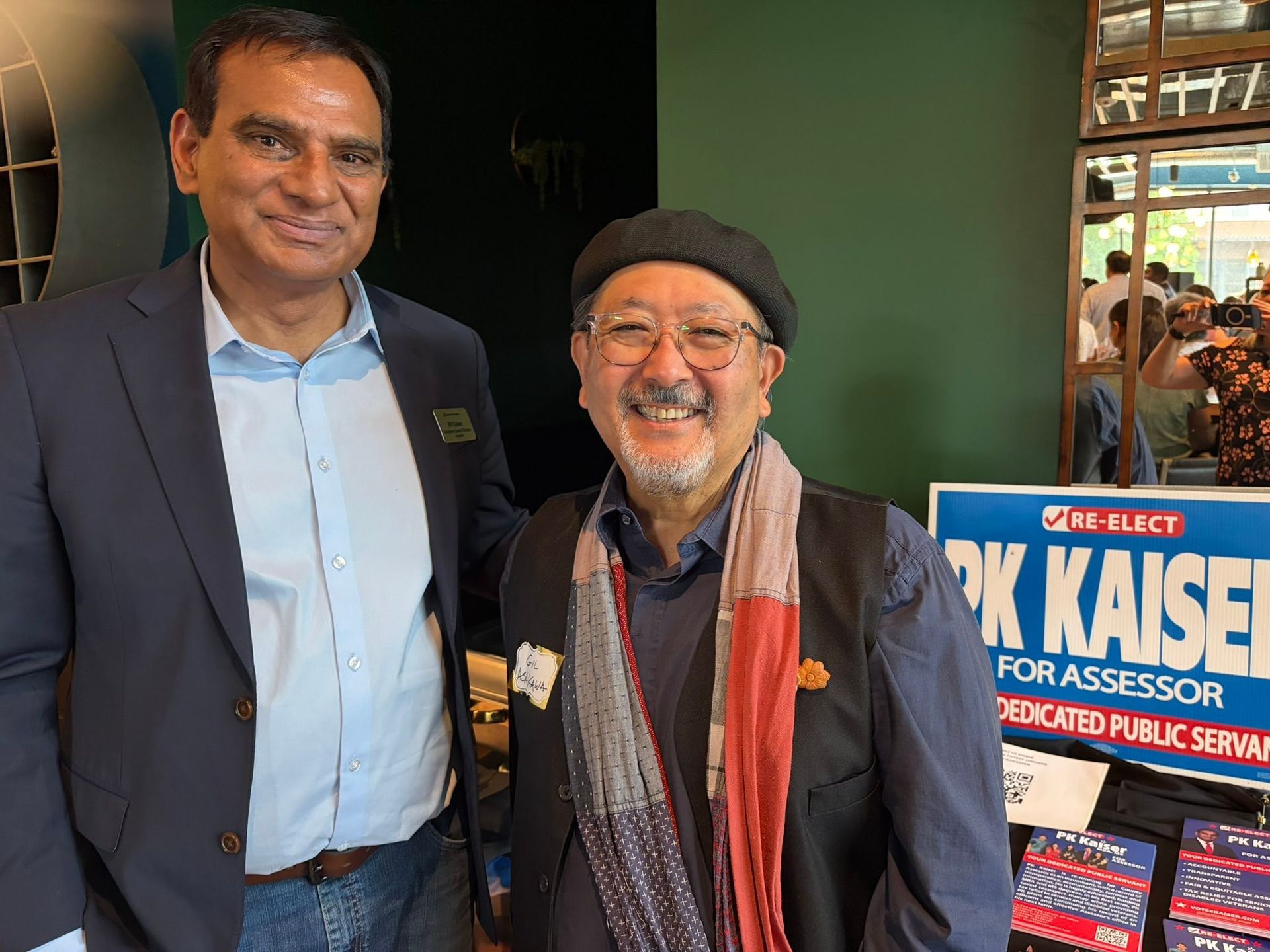 Two smiling men stand indoors beside a blue campaign sign for Rick Kasser.
