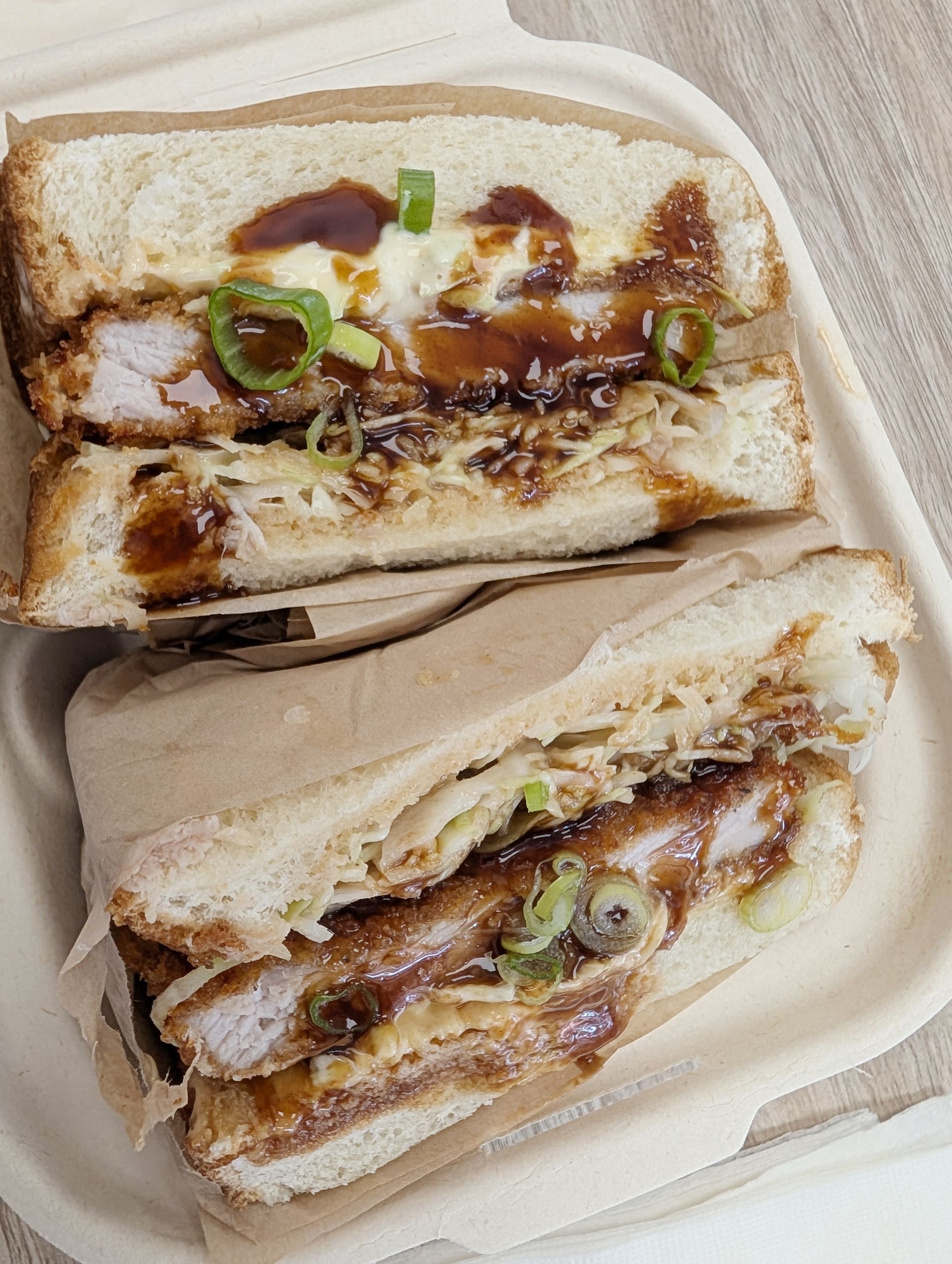 Katsu sandwich cut in half, in a takeout container. Fried pork cutlet, cabbage, sauce, and scallions on toasted bread.