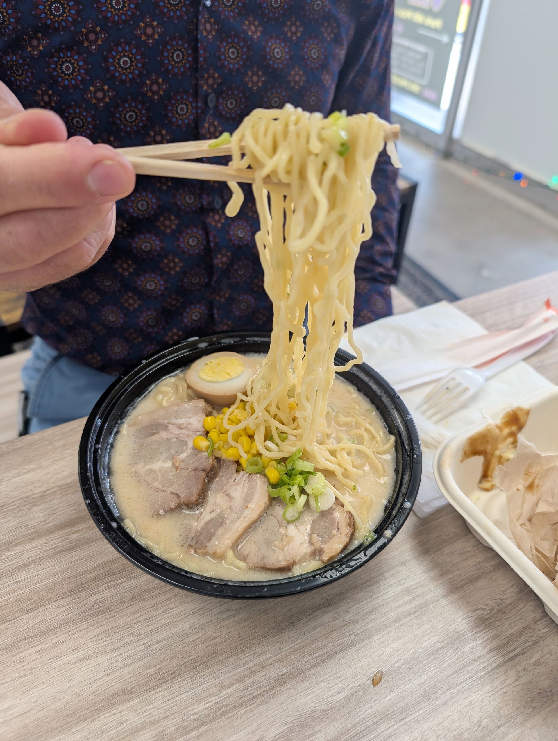 Person eating ramen with chopsticks. Bowl contains noodles, meat, egg, corn, and green onions.