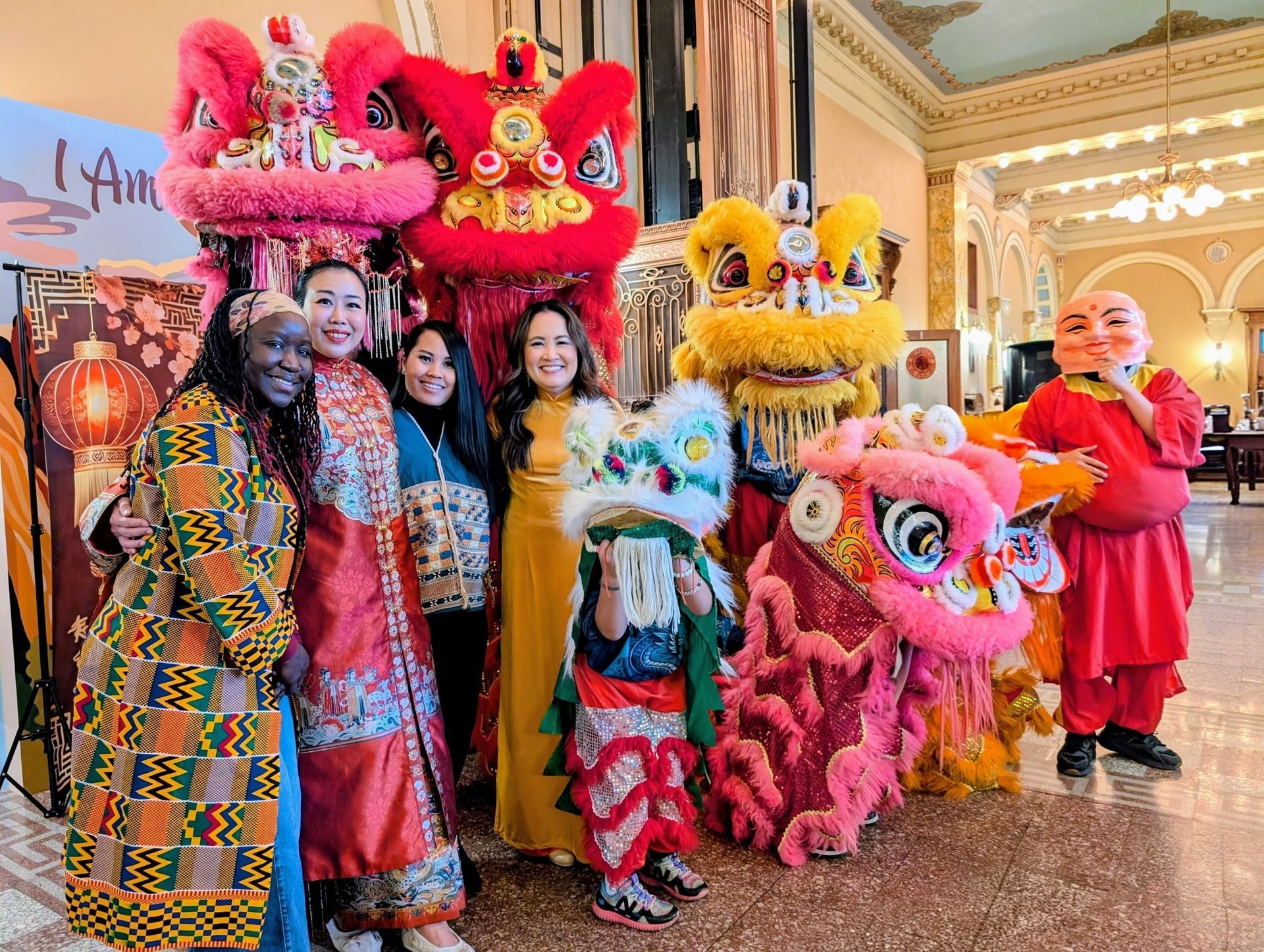 A group poses with colorful Chinese lion dance costumes and a masked performer inside an ornate, warmly lit hall.