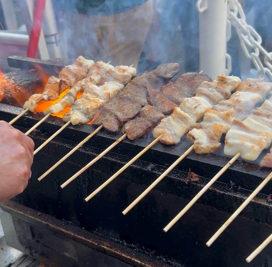 Skewered meats grilling over flames.  A hand turns the skewers.  Outdoor setting, smoke rising.