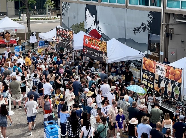 Crowded outdoor food market with white tents, a large mural, and diverse people.