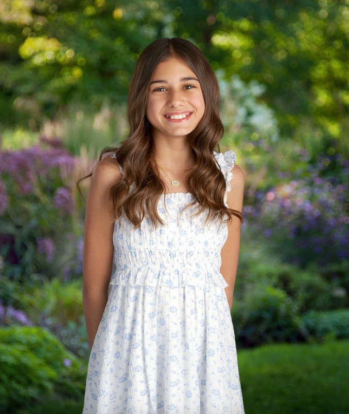 Smiling girl in a white floral dress stands in a garden, arms behind her.