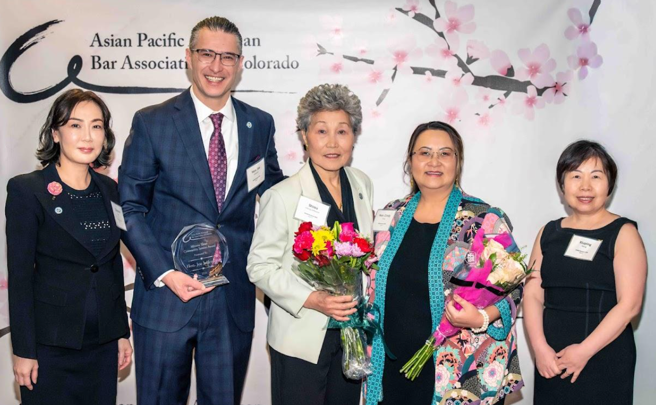 Five people posing at an awards event, with two holding floral bouquets and one holding a glass award.