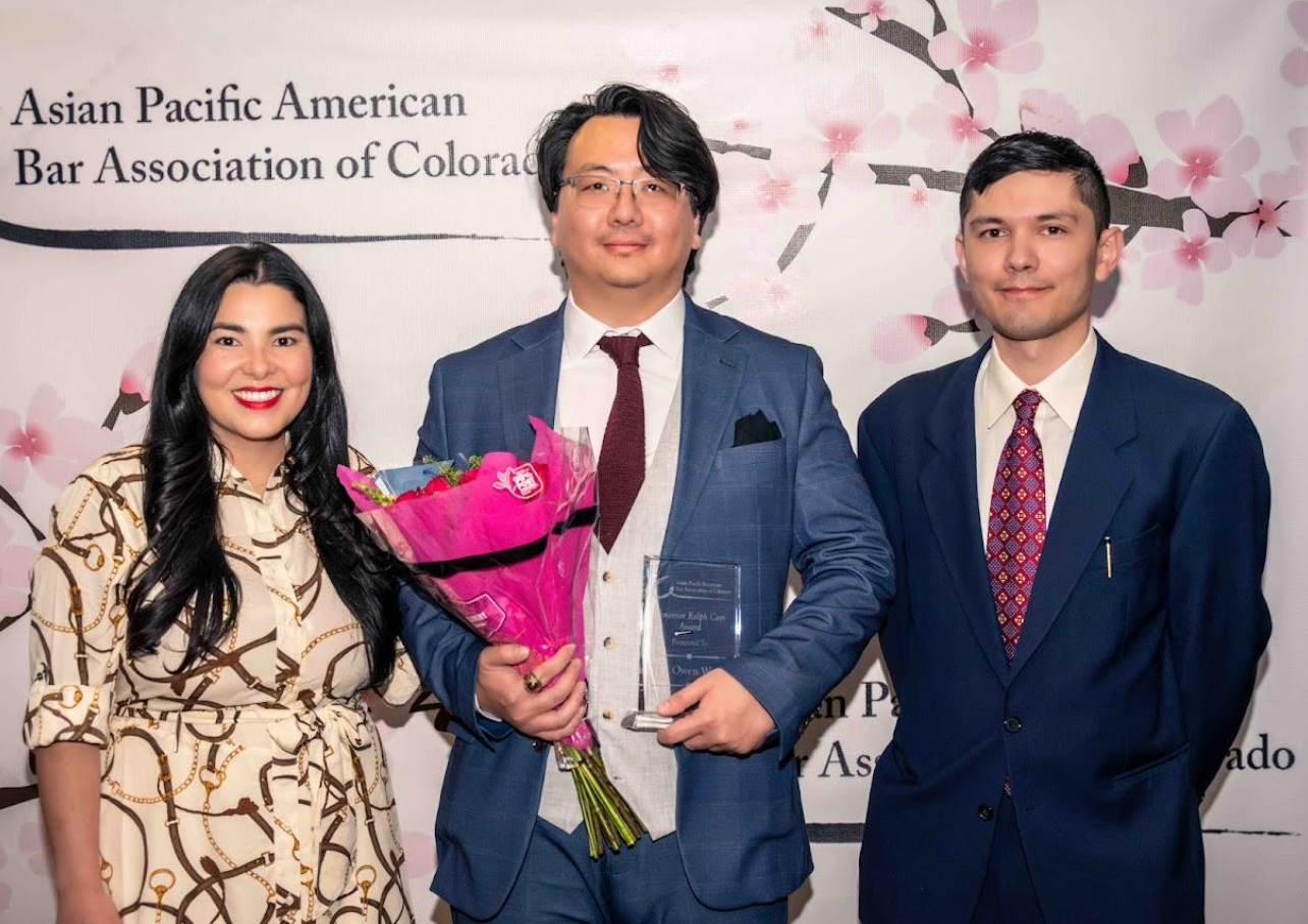 Three people posing at an Asian Pacific American Bar Association of Colorado event, one holding flowers and an award